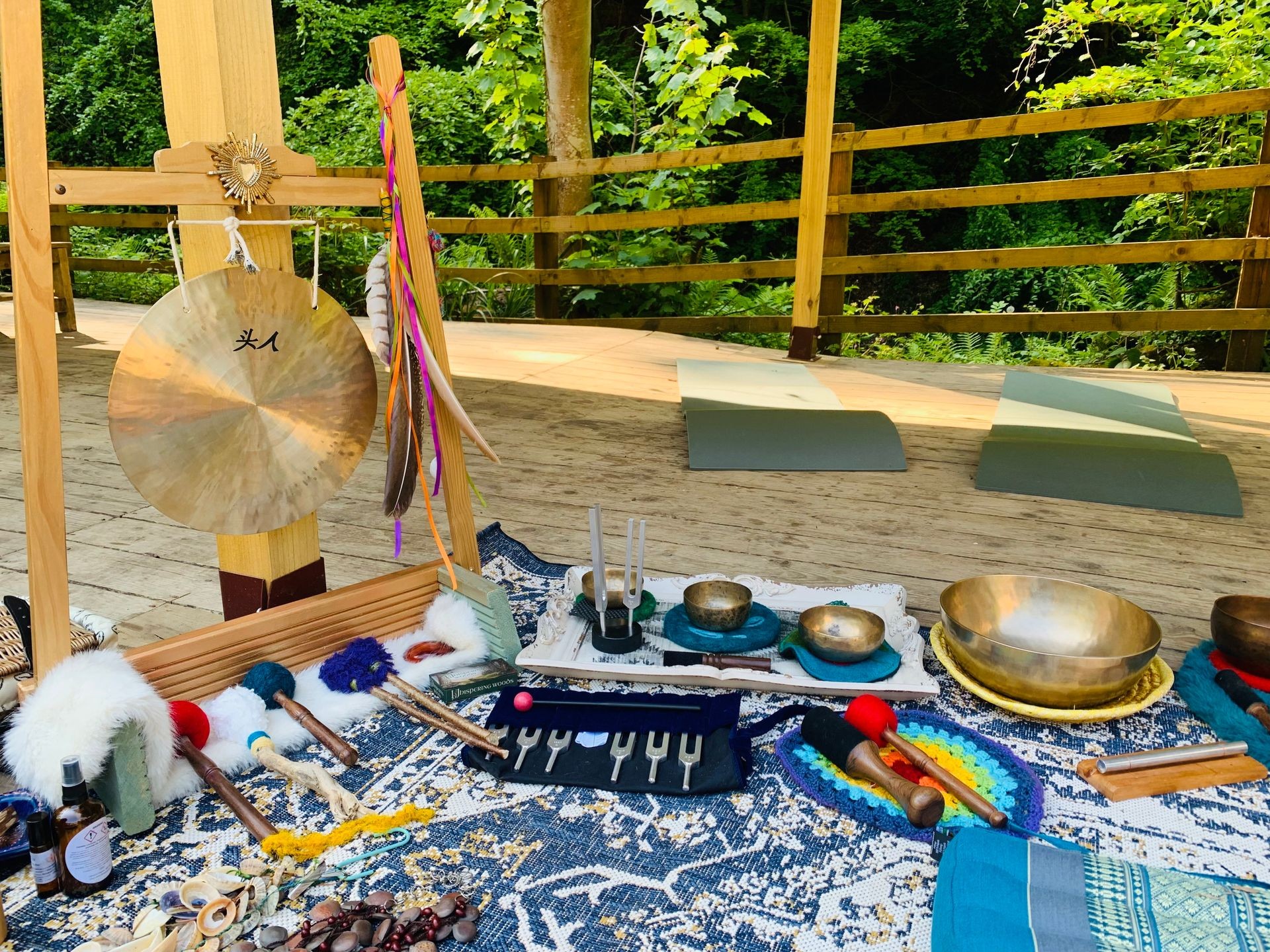 Outdoor setup featuring musical instruments, feathers, and mats on a wooden deck surrounded by greenery.